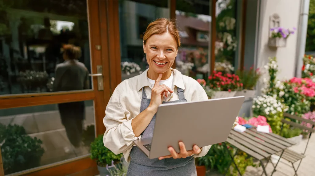 Inhaberin steht vor ihrem Blumenladen mit Laptop in der Hand